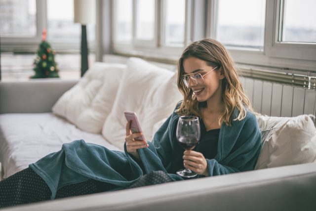 Young woman relaxing in the livingroom drinking wine,and using mobile phone: Aldi offers free wine in exchange or honest reviews