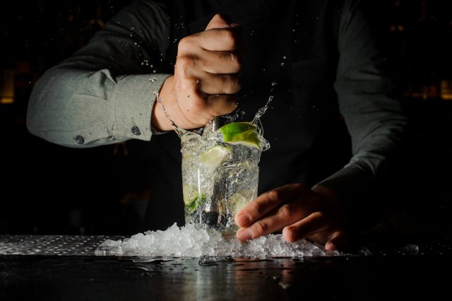 Bartender squeezing juice from fresh lime in a glass using citrus press and splashing it out making an alcoholic cocktail