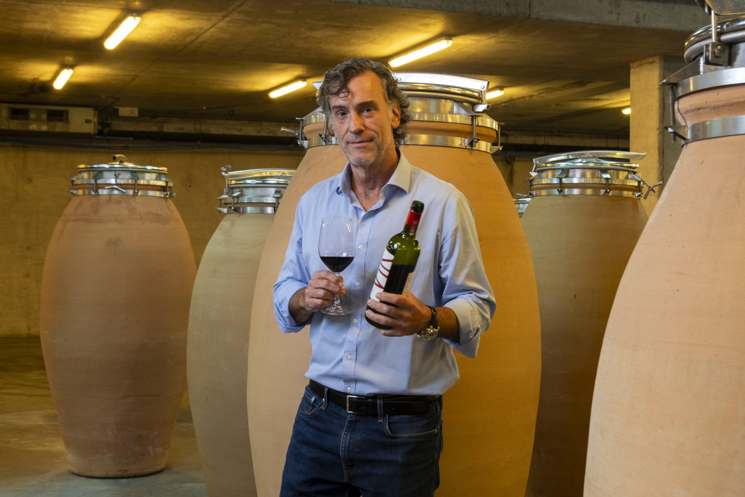 Cristián Vallejo stands in front of clay amphorae, holding an open bottle of Vik wine and a glass of red wine.