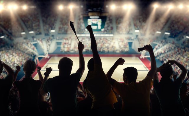 Basketball fans celebrating at a basketball game. We see their silhouettes and fans attributes