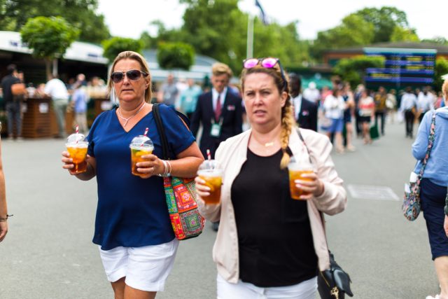 Women carrying plastic cups of Pimm's outdoors, Wimbledon, UK: Whether you’re sipping on Henman Hill or swirling vintage fizz with celebrity tequila moguls in the Royal Box, here’s how to drink like a champion at Wimbledon 2025.