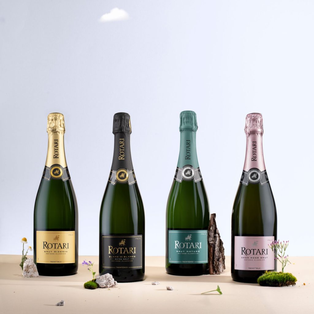 Four bottles of wine - the entire vintage-dated collection of Rotari - sit in front on a plain background, with a few mountain flowers and stones scattered around them.