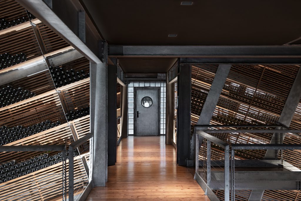 An interior walkway on a bridge leads to a grey door. To either side of the walkway are slanted shelves, filled with bottles of sparkling wine on their sides.