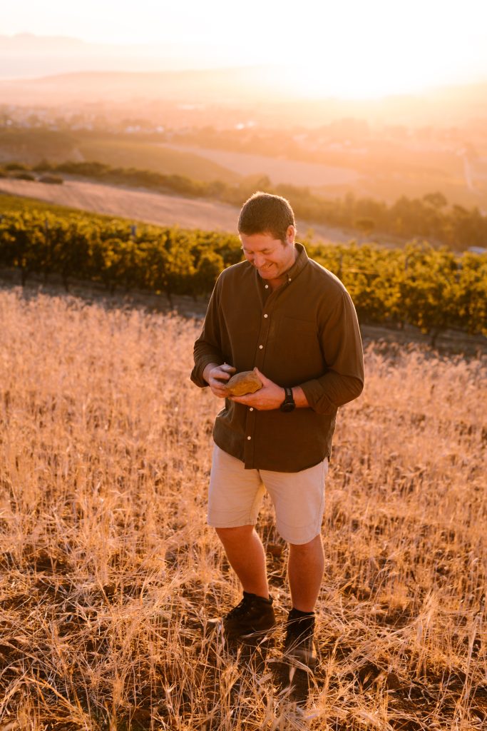 Cellar master Johan Jordaan stands on a loping field, holding a rock, with vineyards in the background.